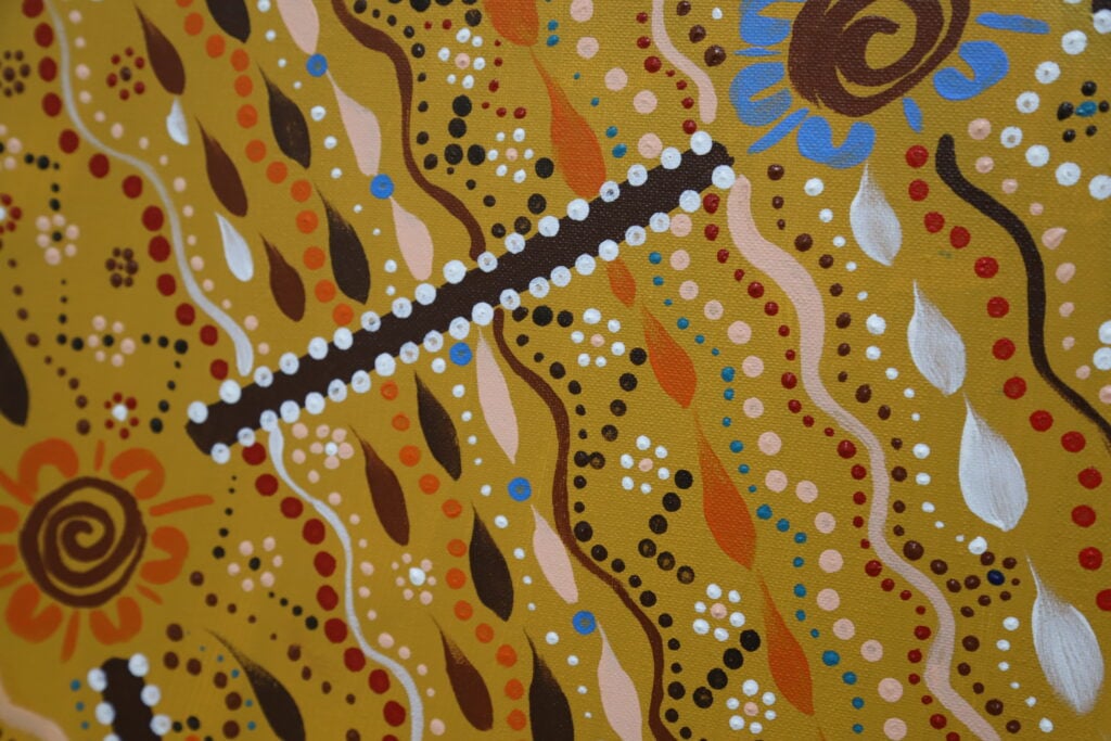 A square canvas with Indigenous art displaying yellow, brown, white, and red dots, wavy lines, and central motif symbols across a mustard-gold background.